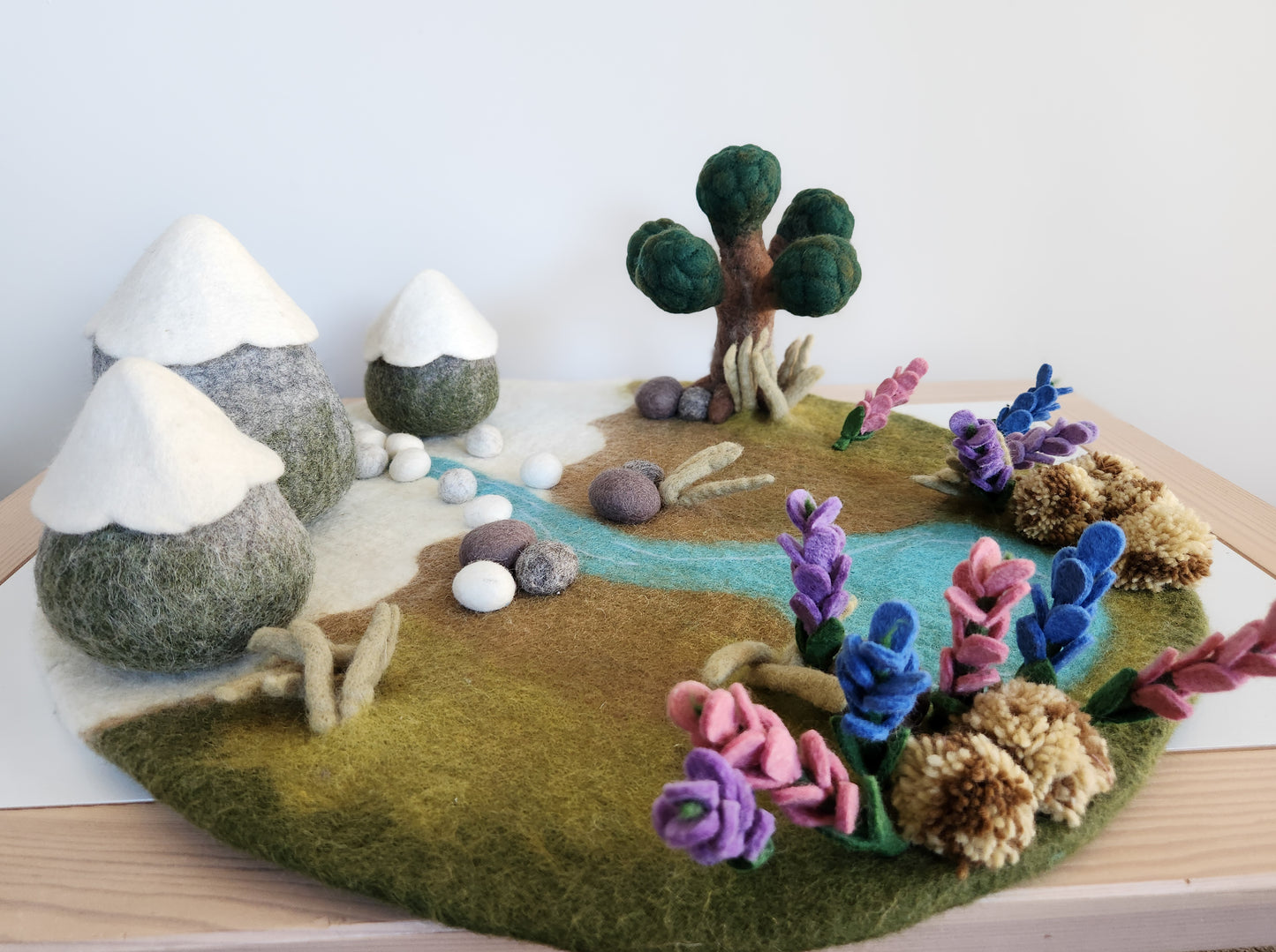 Felted landscape with trees, flowers, and mountains on a wooden surface